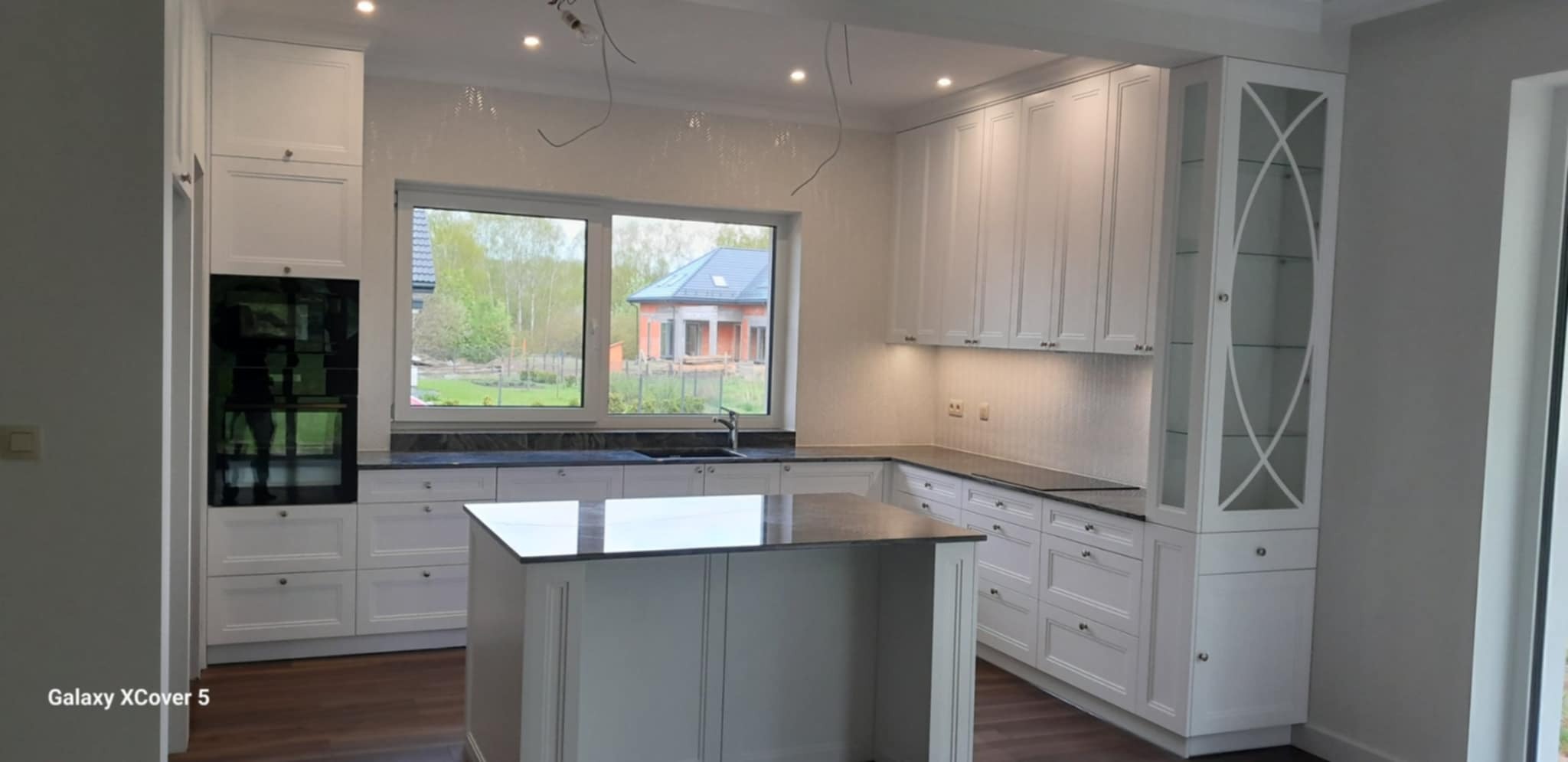 Jasna kuchnia z wyspą i czarnym blatem, widok na dom przez okno. Klasyczne frezowane fronty, oświetlenie podszafkowe. Nowoczesny design z elementami tradycji.