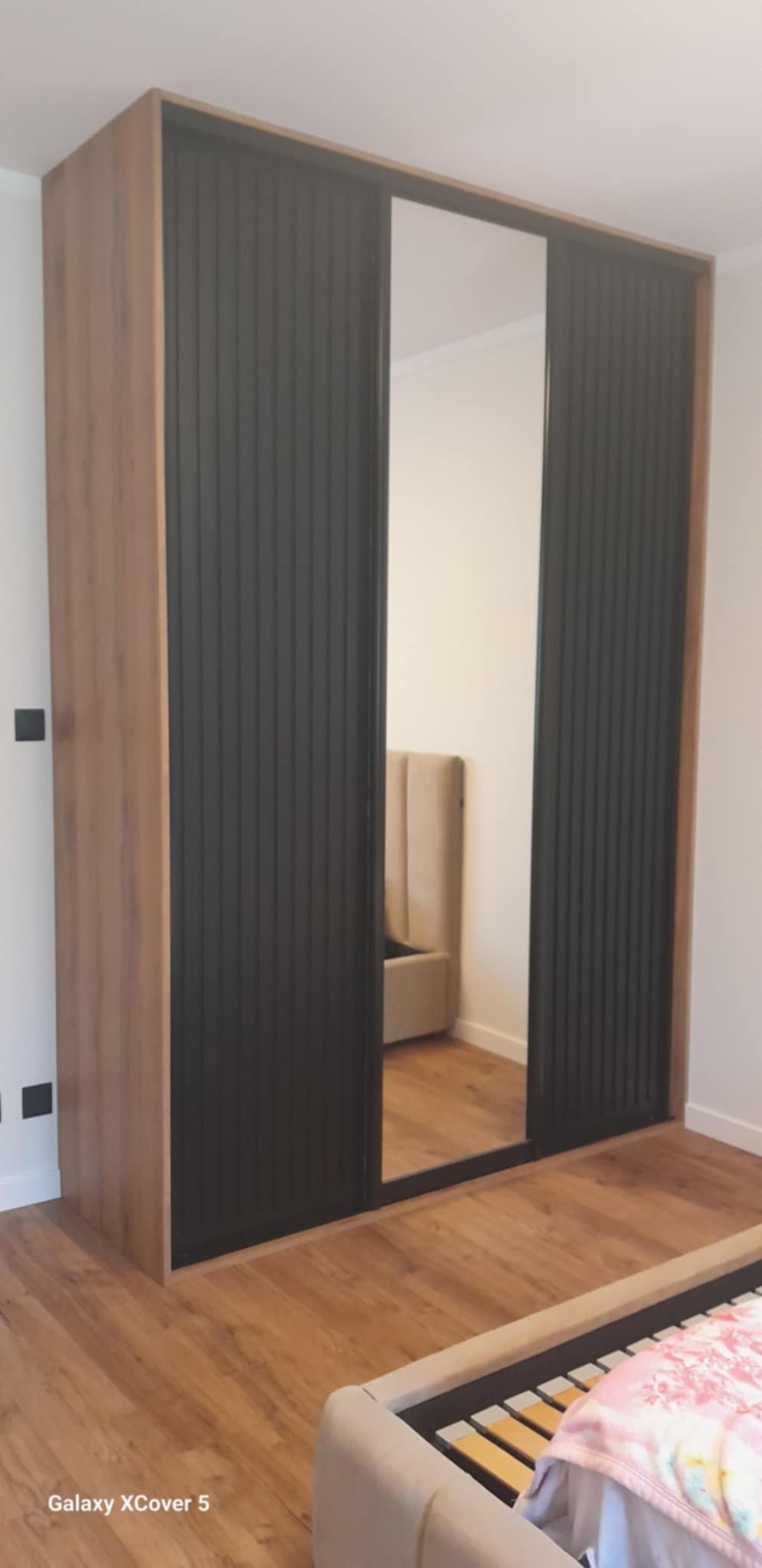 Szafa przesuwna z lustrem i ryflowanymi, czarnymi drzwiami, obudowana drewnopodobną płytą. Widoczny fragment łóżka i podłogi. Minimalistyczny design.