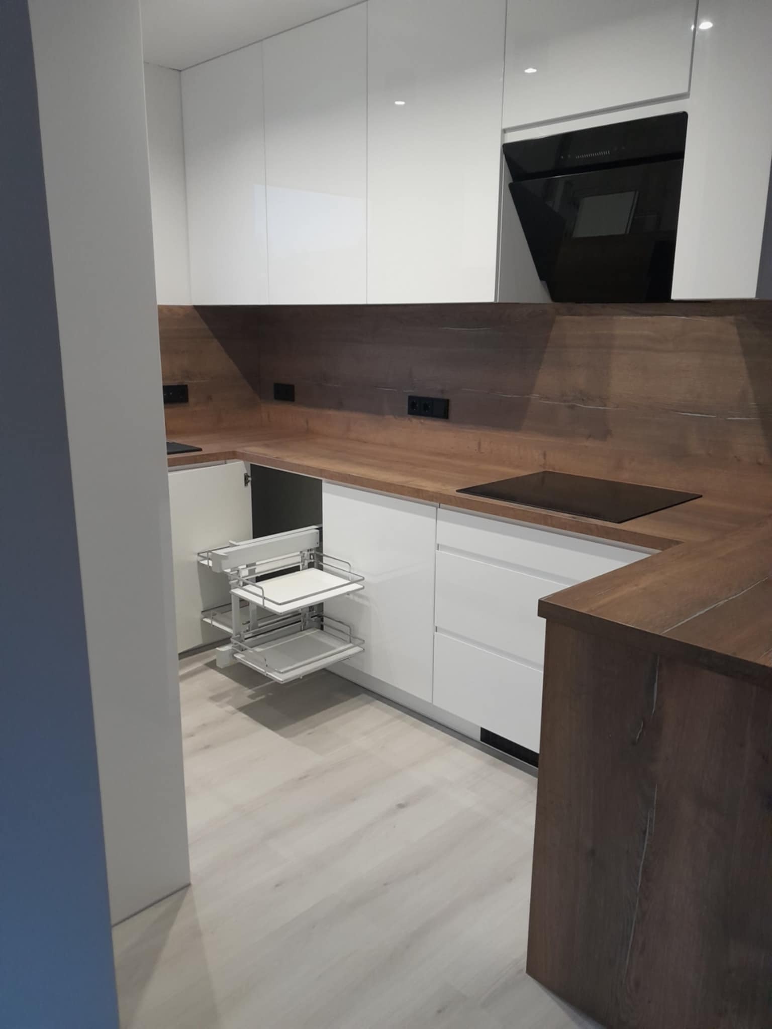 Nowoczesna kuchnia z białymi frontami i drewnianym blatem. Wysuwany kosz cargo w szafce narożnej. Czarny okap nad płytą grzewczą. Jasna podłoga.