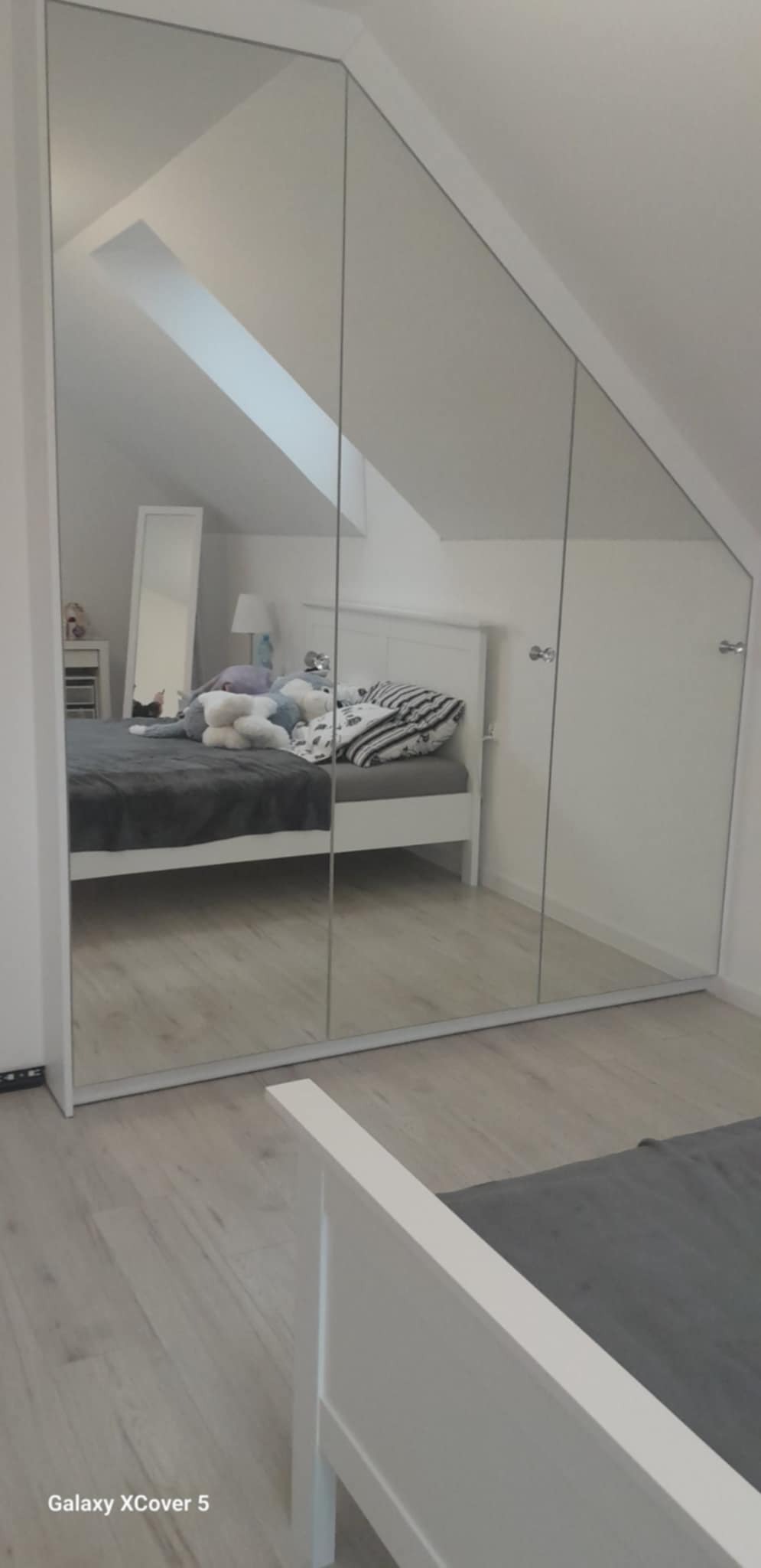 Biała szafa wnękowa z lustrzanymi drzwiami przesuwnymi, w pokoju na poddaszu. Widoczne odbicie łóżka i okna dachowego. Minimalistyczny design.
