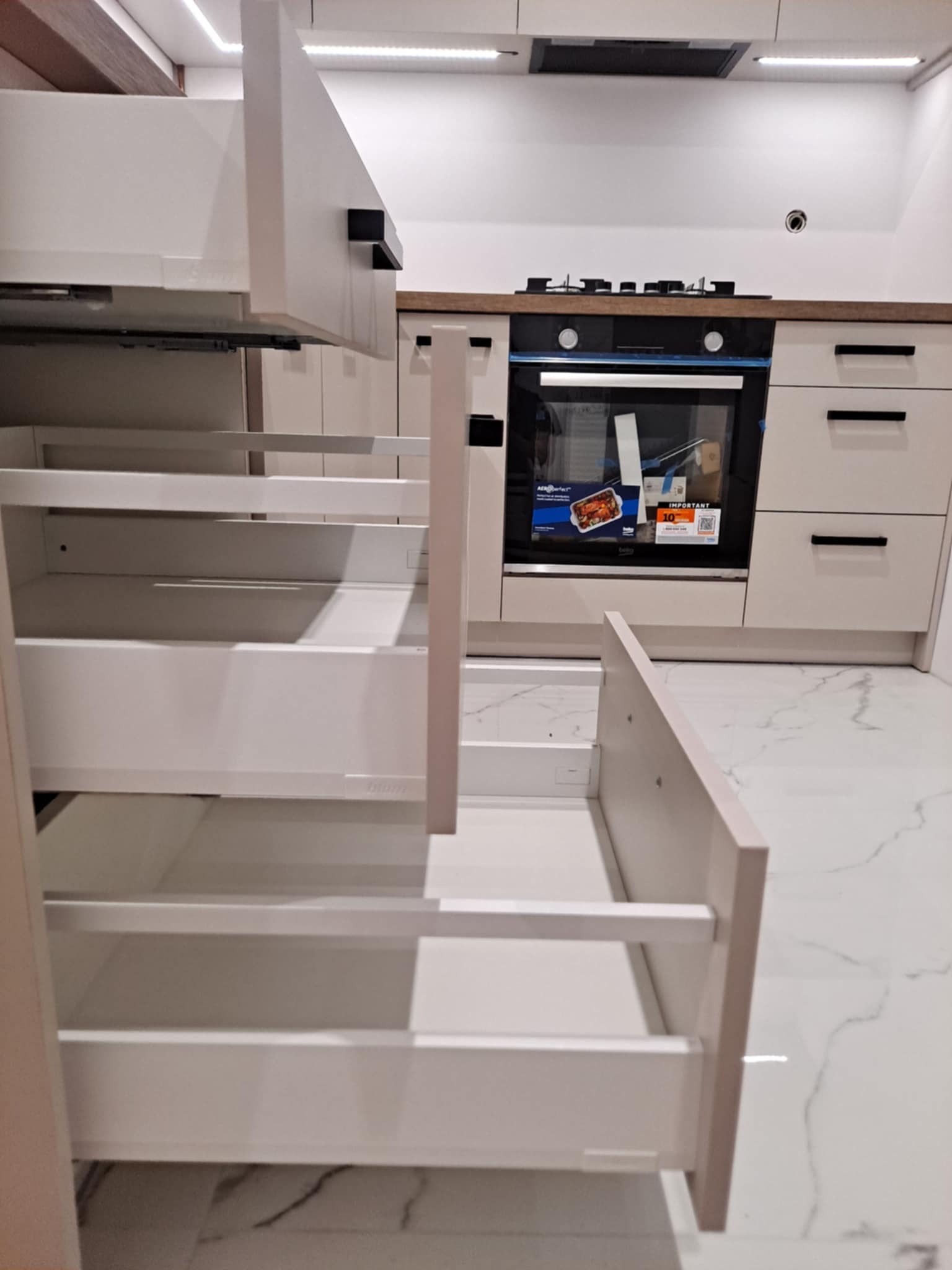 Jasna kuchnia na wymiar z wysuwanymi szufladami cargo. Widoczne czarne, minimalistyczne uchwyty i marmurowa podłoga. Oświetlenie LED podszafkowe.