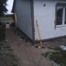 Docieplenia elewacje - Budynek w trakcie ocieplania, widoczny styropian na ścianach, poziomica oparta o ścianę, okno z parapetem i przygotowane materiały budowlane obok budynku.