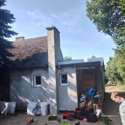 Docieplenia elewacje - Dom jednorodzinny z szarą elewacją w trakcie remontu, z widocznymi materiałami izolacyjnymi obok i piaskownicą z zabawkami na pierwszym planie, z chłopcem przyglądającym się.