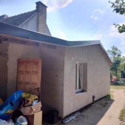 Docieplenia elewacje - Wykończona elewacja budynku jednokondygnacyjnego z oknem PCV, widoczny fragment dachu i komina sąsiedniego budynku, obok wejścia bałagan.