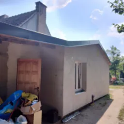 Wykończona elewacja budynku jednokondygnacyjnego z oknem PCV, widoczny fragment dachu i komina sąsiedniego budynku, obok wejścia bałagan.