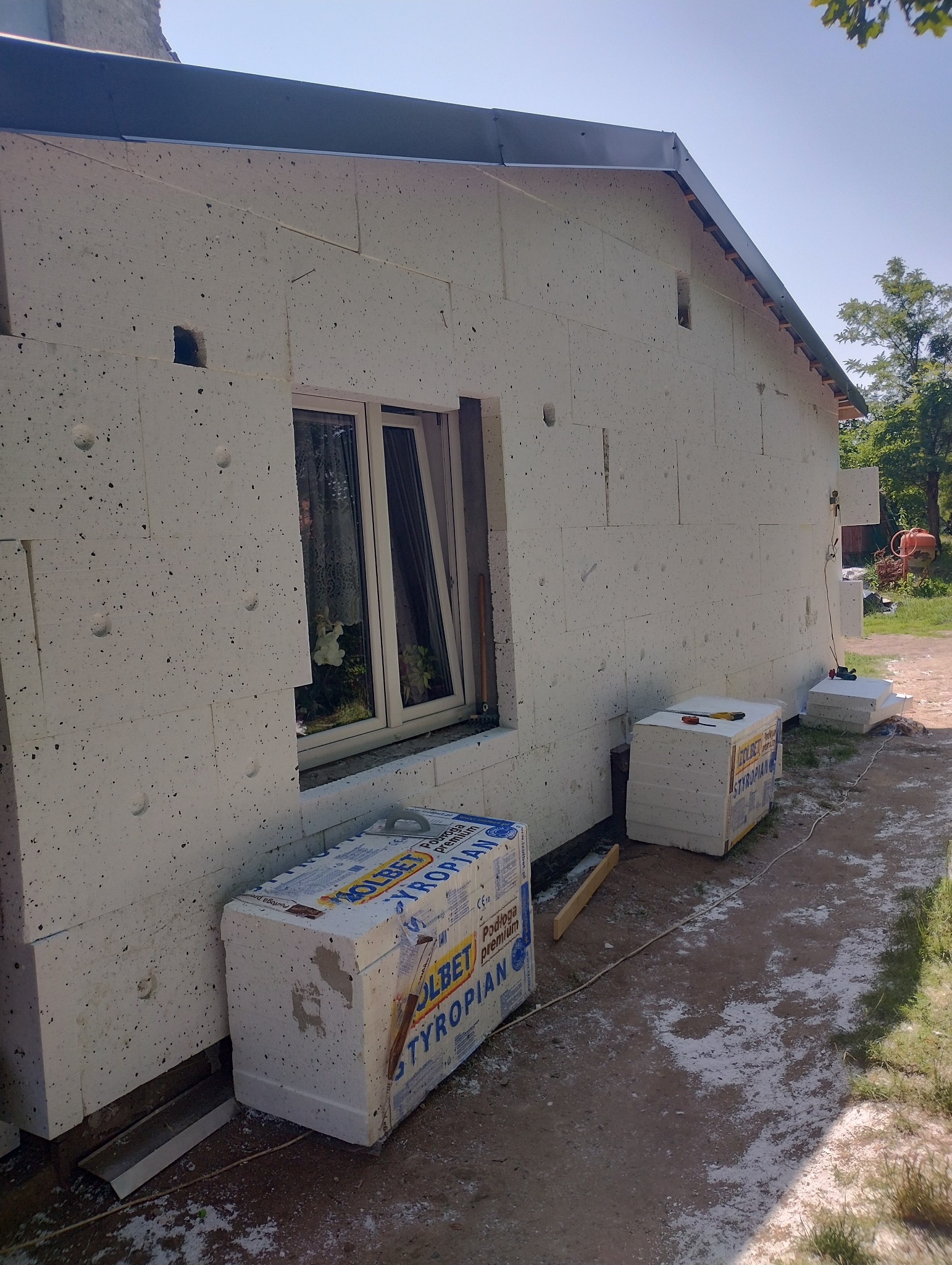 Dom jednorodzinny w trakcie prac związanych z izolacją termiczną ścian zewnętrznych, widoczne płyty styropianowe, okno z firanką oraz opakowania styropianu marki Izolbet.