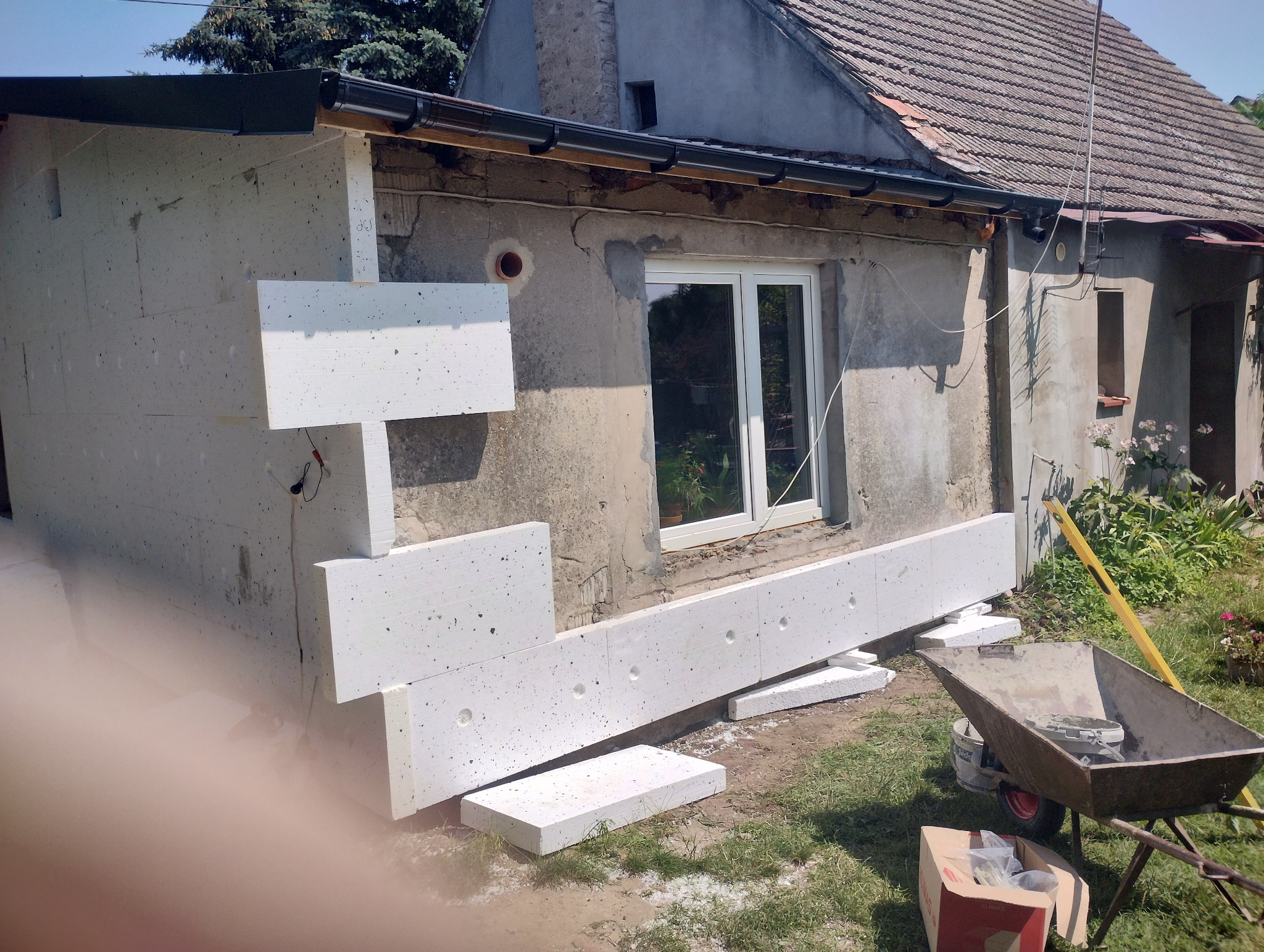 Fragment ściany domu z widocznymi etapami montażu białych płyt izolacyjnych, z przodu taczka z narzędziami, w tle budynek z oknem.