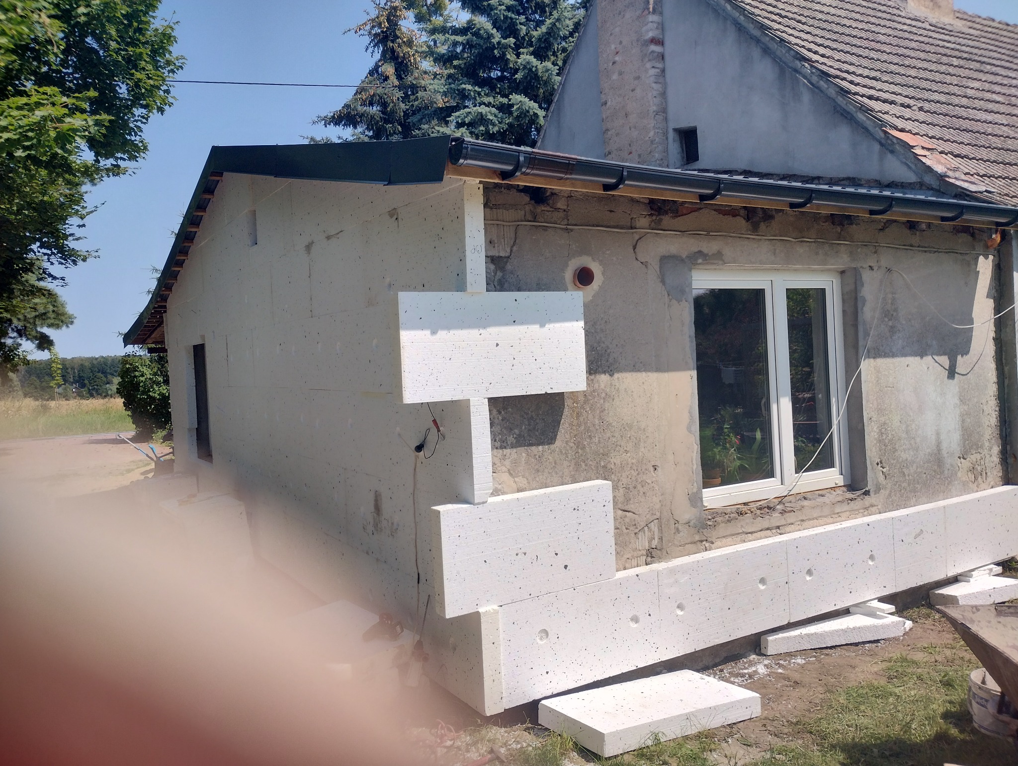 Ocieplanie elewacji starego domu z widocznymi fragmentami styropianu, zamontowanymi na szarej, nierównej powierzchni ściany, z nowym oknem i elementami orynnowania.