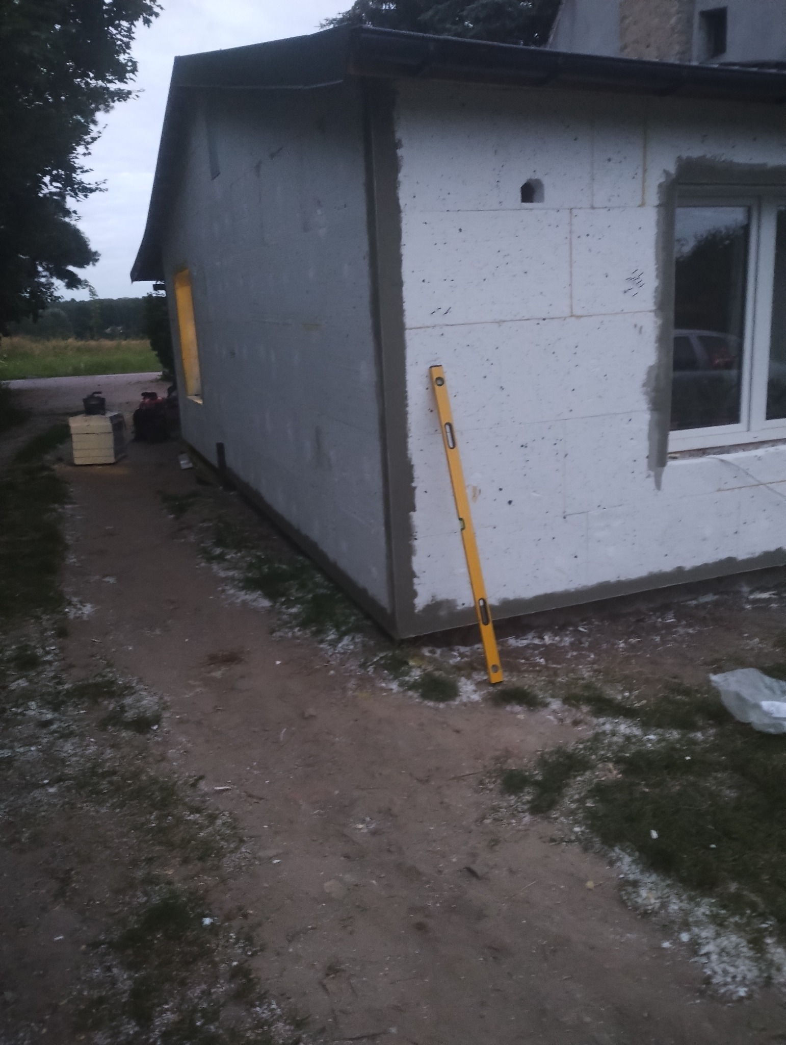 Budynek w trakcie ocieplania, widoczny styropian na ścianach, poziomica oparta o ścianę, okno z parapetem i przygotowane materiały budowlane obok budynku.
