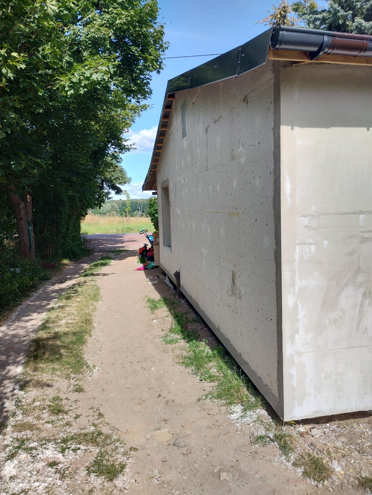 Ściana budynku w trakcie ocieplania, widoczne płyty izolacyjne i warstwa szpachli, obok ścieżka gruntowa z zielenią, w tle zabawki dziecięce.