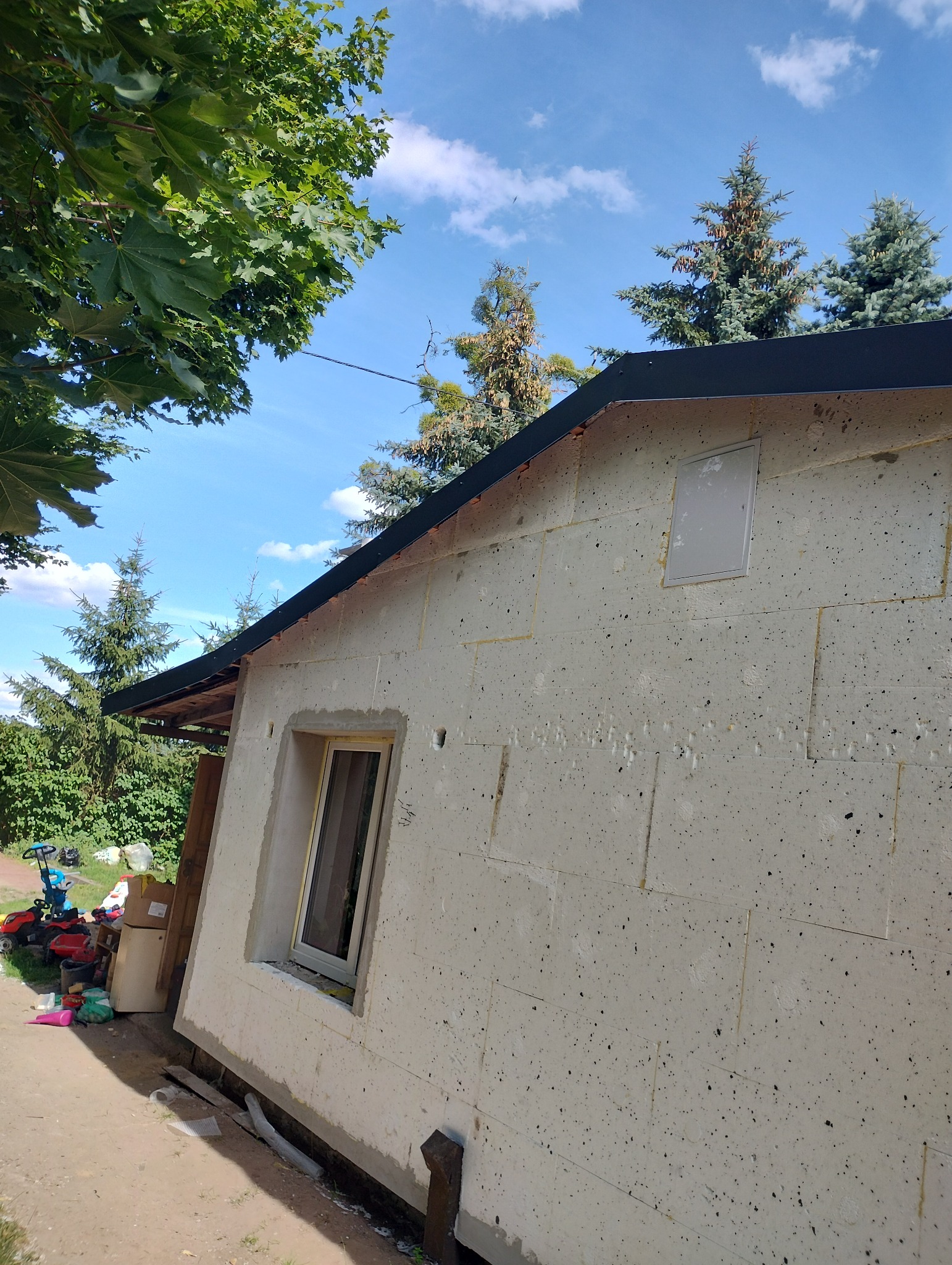 Budynek w trakcie ocieplania elewacji, widoczne płyty styropianowe z czarnymi plamkami, zamontowane okno oraz czarny dach, w tle zieleń drzew i błękitne niebo.