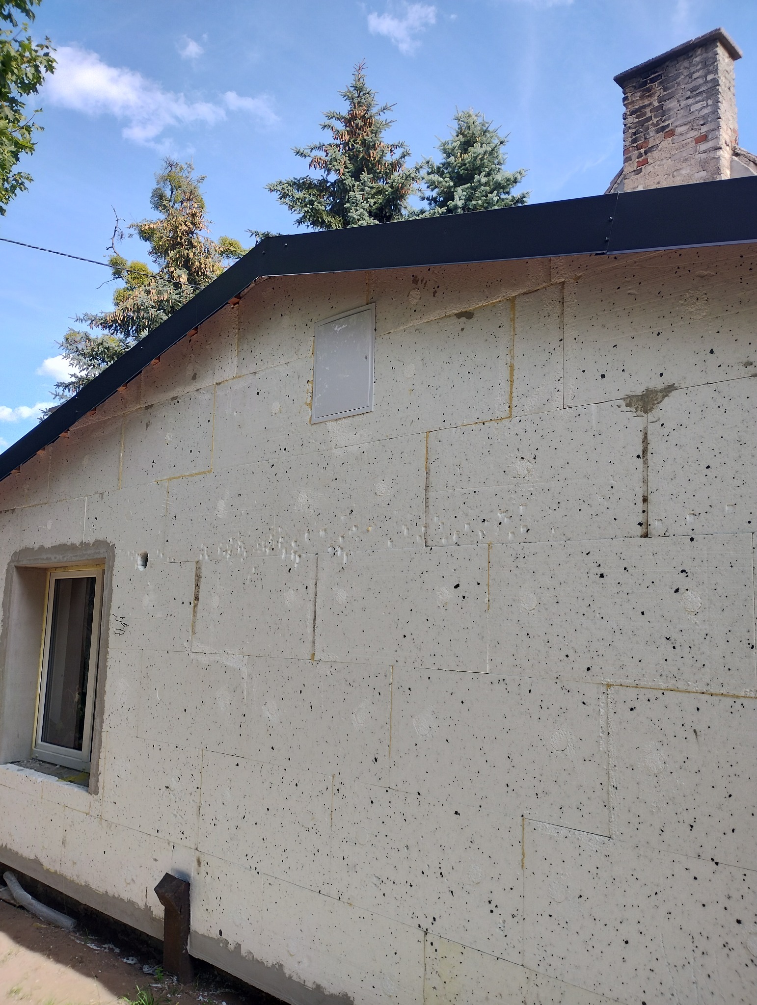 Ściana budynku w trakcie prac, pokryta płytami izolacyjnymi z widocznym oknem i wentylacją, przygotowana do dalszych etapów wykończenia.