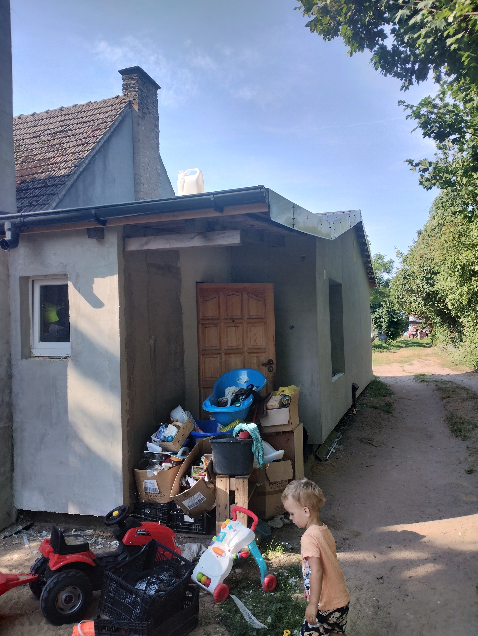 Surowy budynek w trakcie prac wykończeniowych elewacji, z oknami bez szyb, zabałaganiony teren budowy z zabawkami dziecięcymi. Nowa elewacja w jasnym kolorze kontrastuje z ciemnym dachem i zielenią...