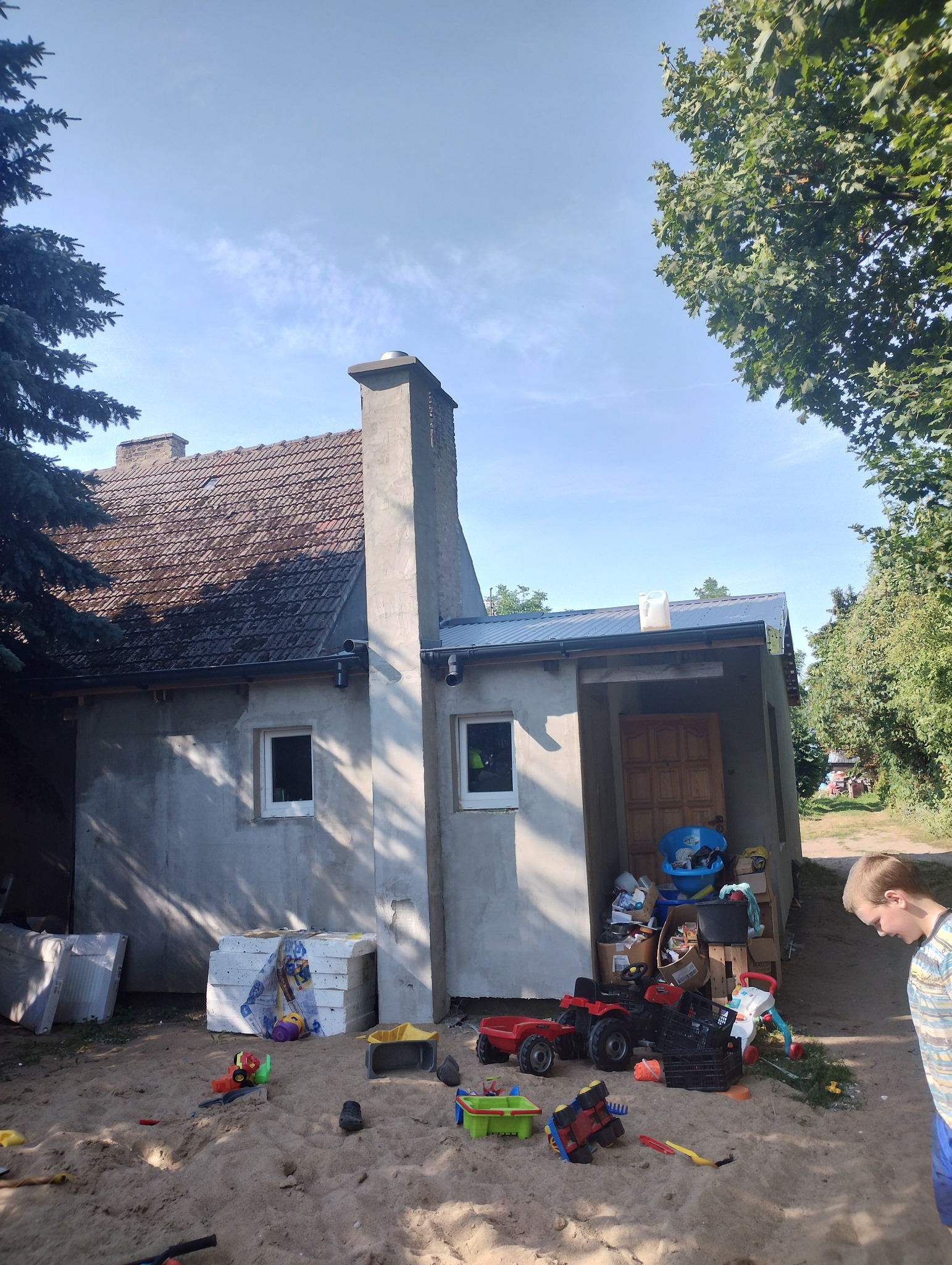 Dom jednorodzinny z szarą elewacją w trakcie remontu, z widocznymi materiałami izolacyjnymi obok i piaskownicą z zabawkami na pierwszym planie, z chłopcem przyglądającym się.