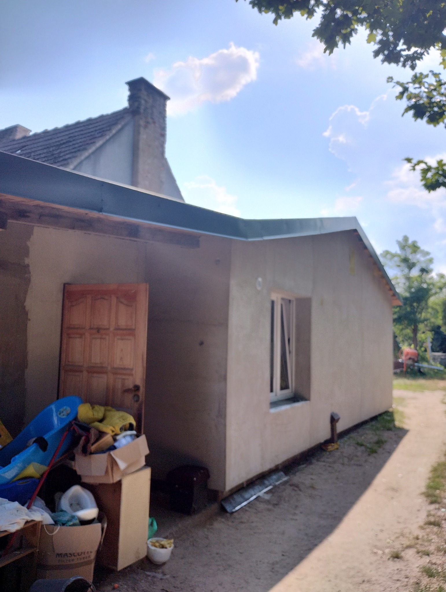Wykończona elewacja budynku jednokondygnacyjnego z oknem PCV, widoczny fragment dachu i komina sąsiedniego budynku, obok wejścia bałagan.