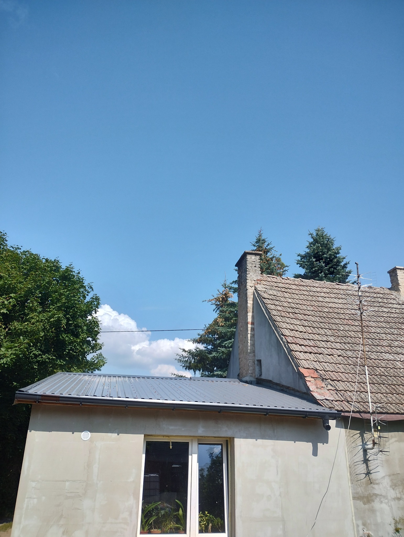 Surowa elewacja budynku z nowym metalowym dachem, widok na komin z cegły na tle błękitnego nieba i drzew.