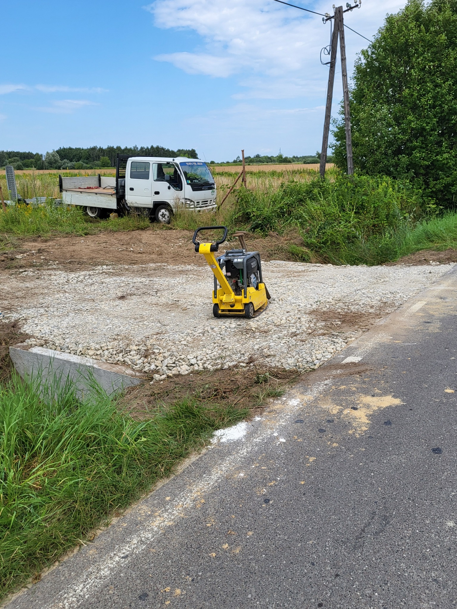Żółta zagęszczarka Wacker Neuson stoi na warstwie tłucznia, obok widać betonową konstrukcję i białą ciężarówkę na tle pola i drzew.
