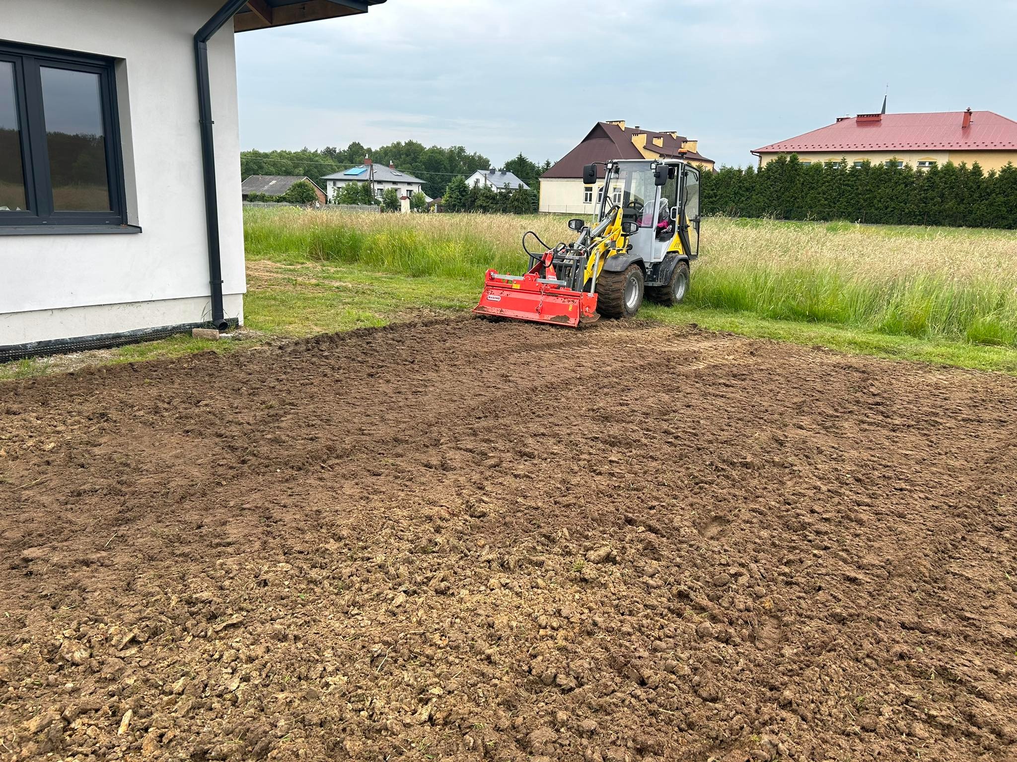 Mały traktor z glebogryzarką przygotowuje ziemię pod trawnik na działce przy domu jednorodzinnym.