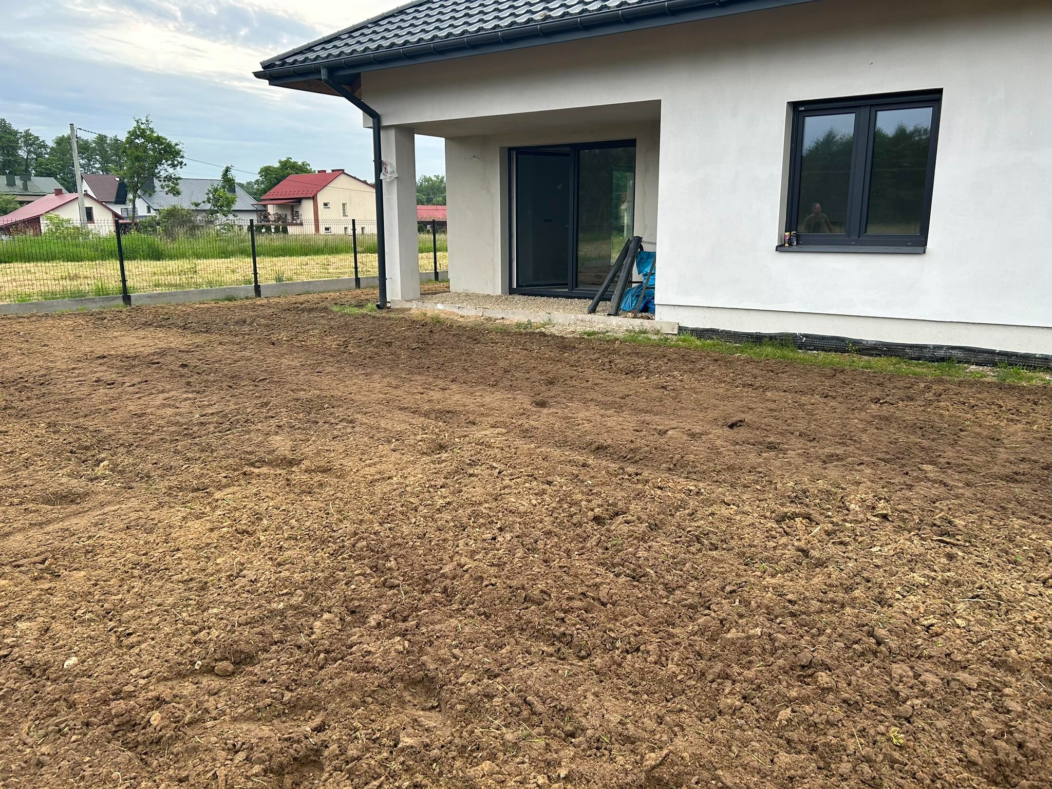 Przygotowany teren pod trawnik przy nowo wybudowanym domu z widoczną czarną folią izolacyjną przy fundamencie.