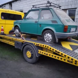 Zielony Fiat 126p z bagażnikiem dachowym, zabezpieczony pasami, transportowany na żółtej lawecie z numerem telefonu.