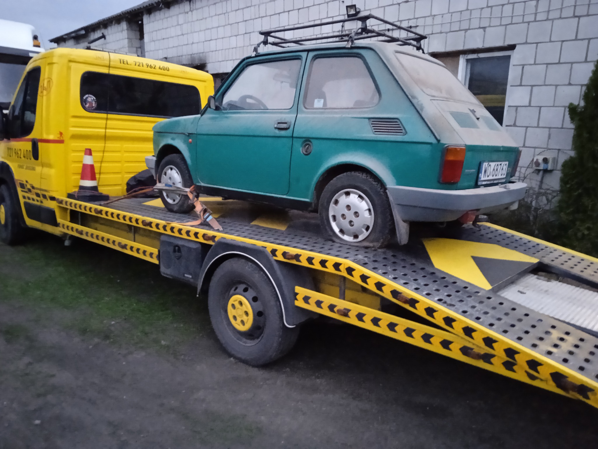 Zielony Fiat 126p z bagażnikiem dachowym, zabezpieczony pasami, transportowany na żółtej lawecie z numerem telefonu.