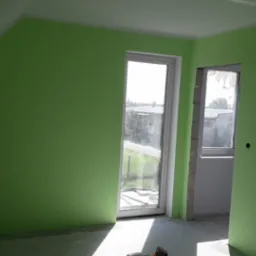 Świeżo pomalowane na jasnozielono wnętrze pokoju z oknem i otwartym przejściem, widoczne gniazdka elektryczne bez osprzętu, surowy stan wykończenia podłogi i ościeżnicy.