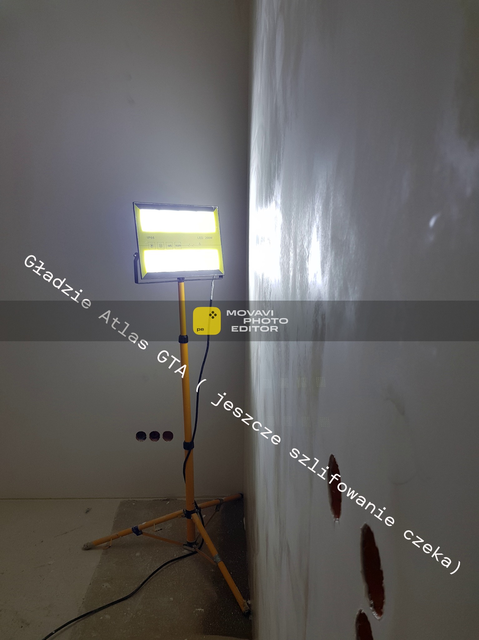 Ściana po nałożeniu gładzi Atlas GTA, oświetlona lampą budowlaną. Widoczne niedoskonałości wymagające szlifowania. Przygotowanie do malowania.