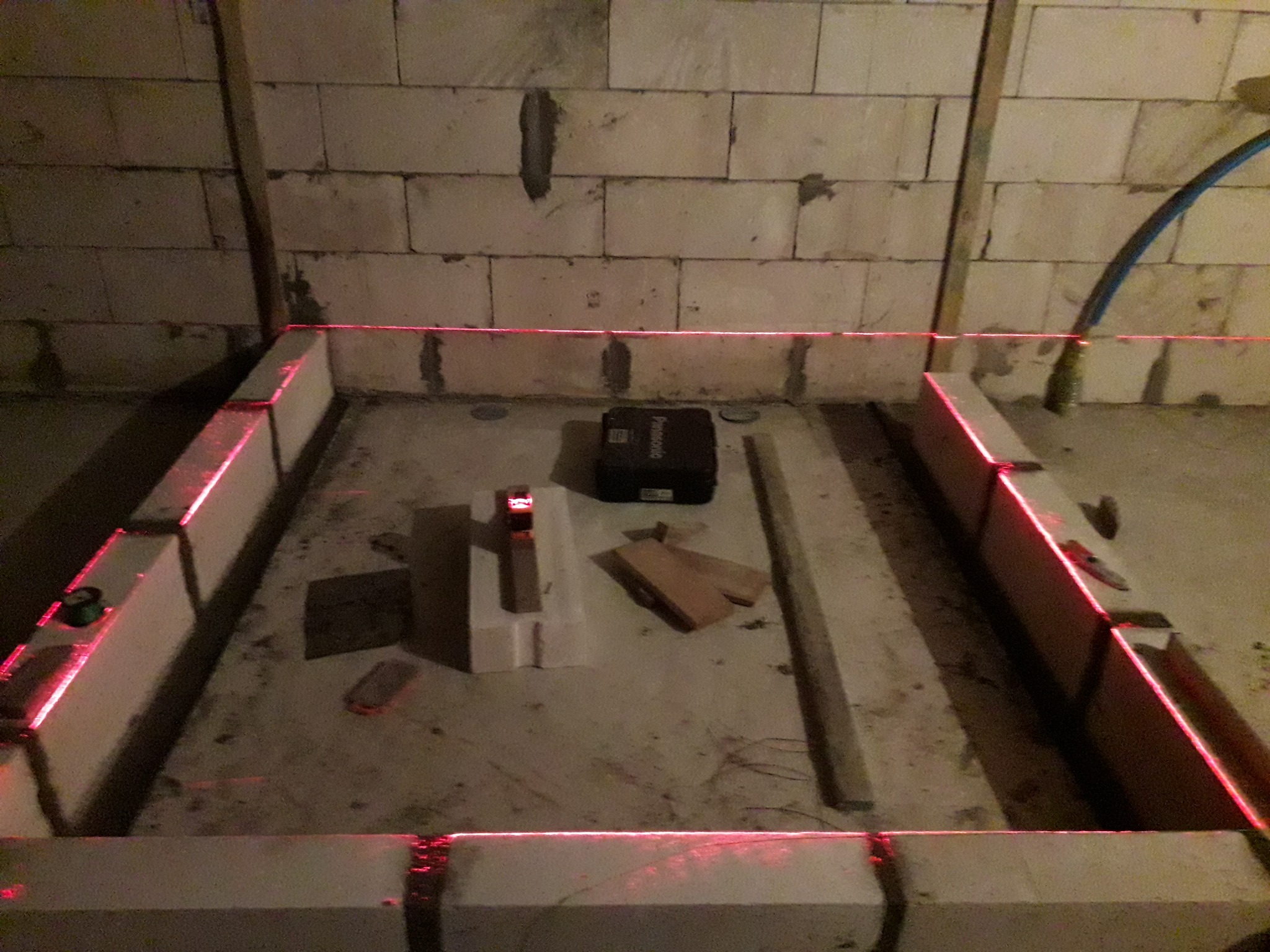 Przygotowanie podłoża pod wylewkę: bloczki betonowe ustawione w kwadrat, wyznaczanie poziomu za pomocą lasera krzyżowego, widoczna skrzynka na narzędzia i deski na podłodze.
