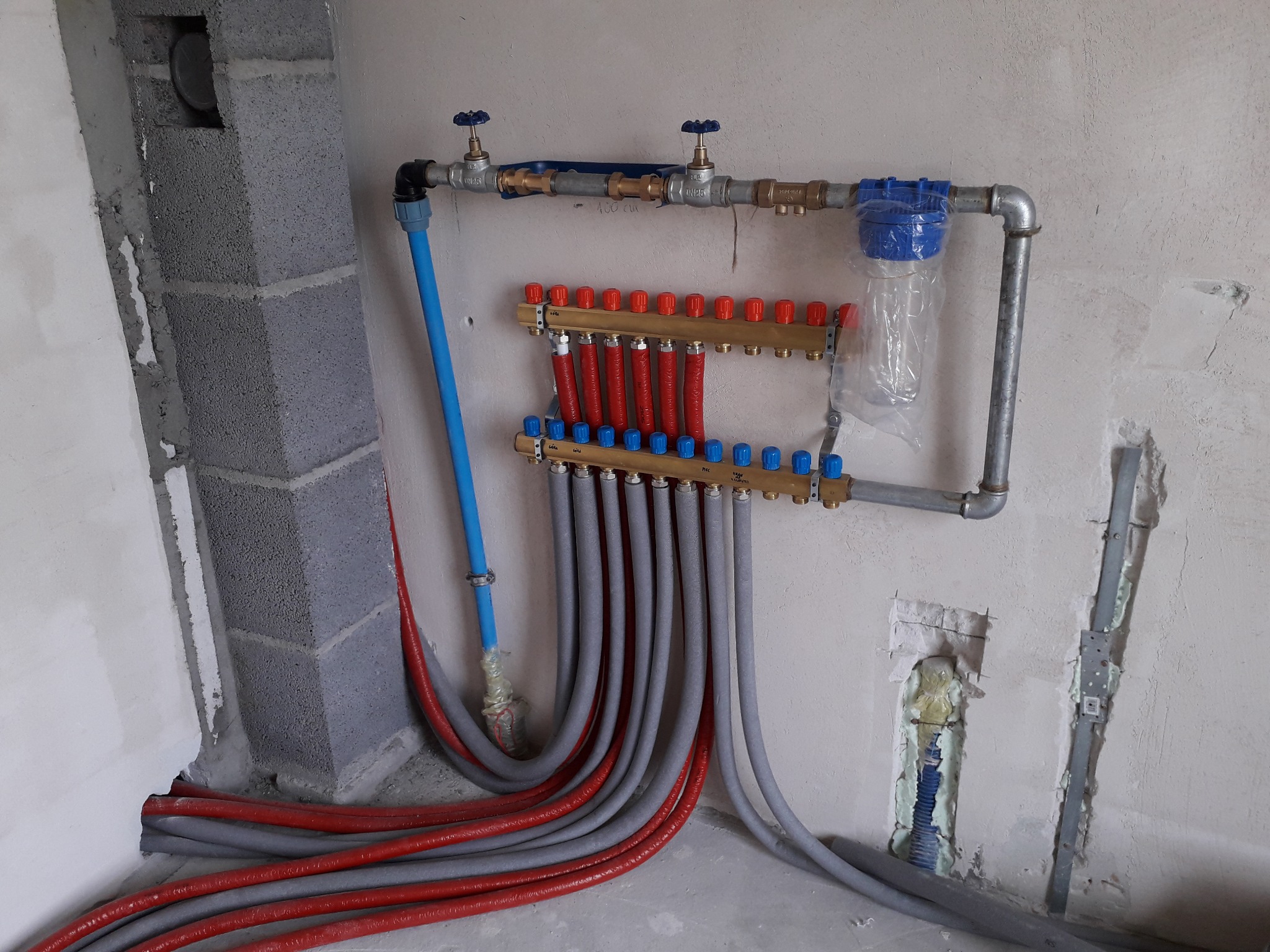 Rozdzielacz ogrzewania podłogowego z czerwonymi i szarymi rurami w surowym stanie, widoczne niedokończone ściany i fragment instalacji wodnej.