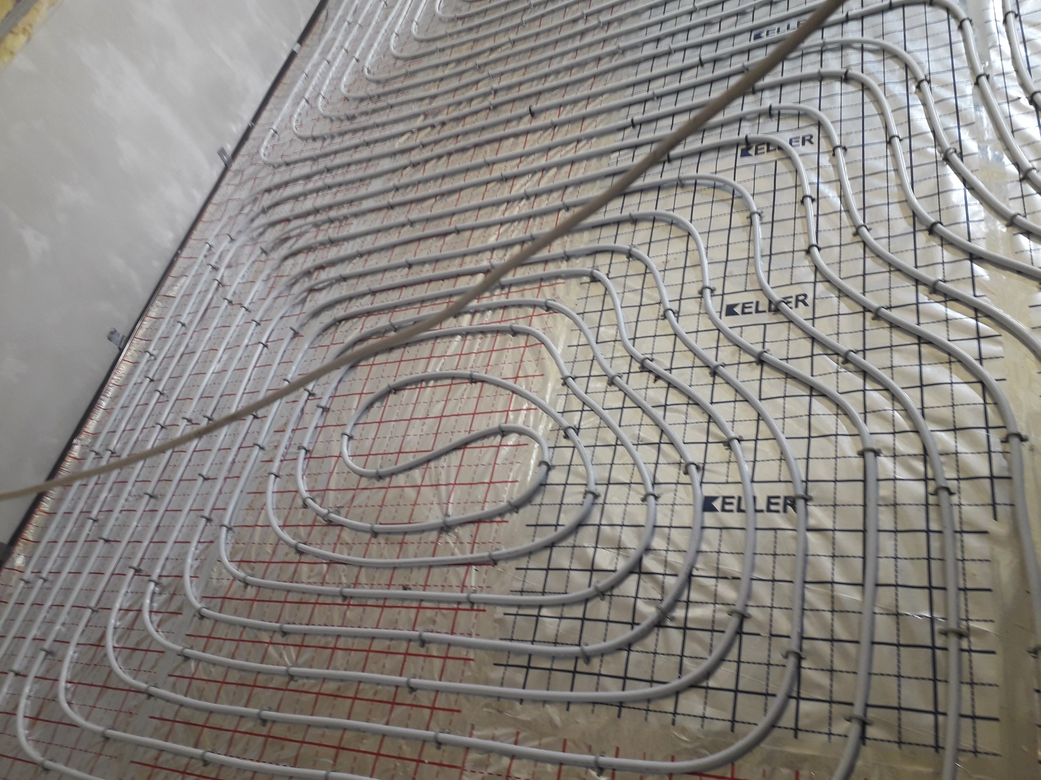 Układanie rur ogrzewania podłogowego na folii z siatką, widoczne mocowania i fragment ściany w surowym stanie.