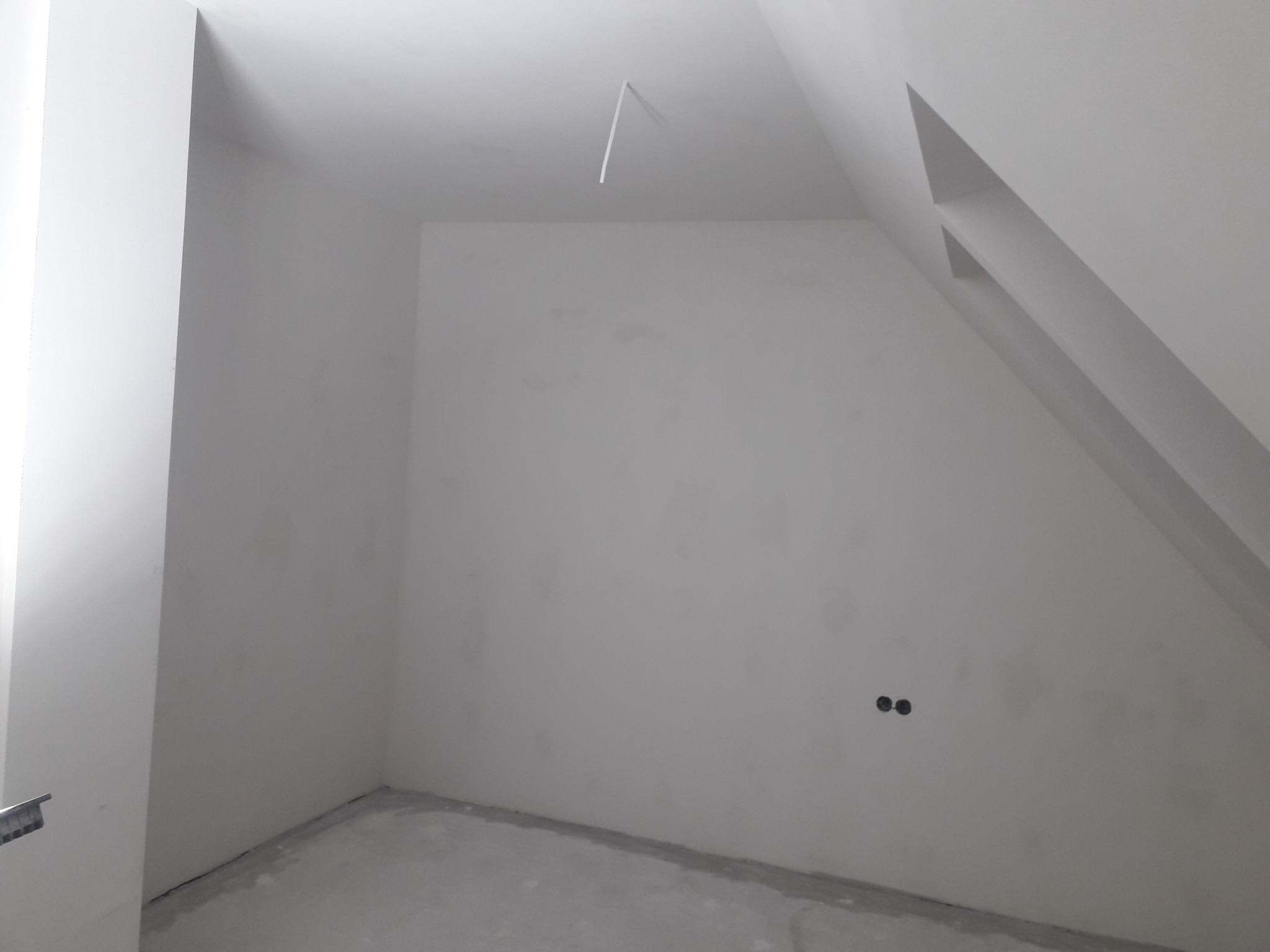 Wnętrze pokoju na poddaszu w Szamocinie przed malowaniem, widoczne gładkie ściany przygotowane do dalszych prac wykończeniowych, dwa otwory na gniazdka elektryczne i wystający kabel ze sufitu.