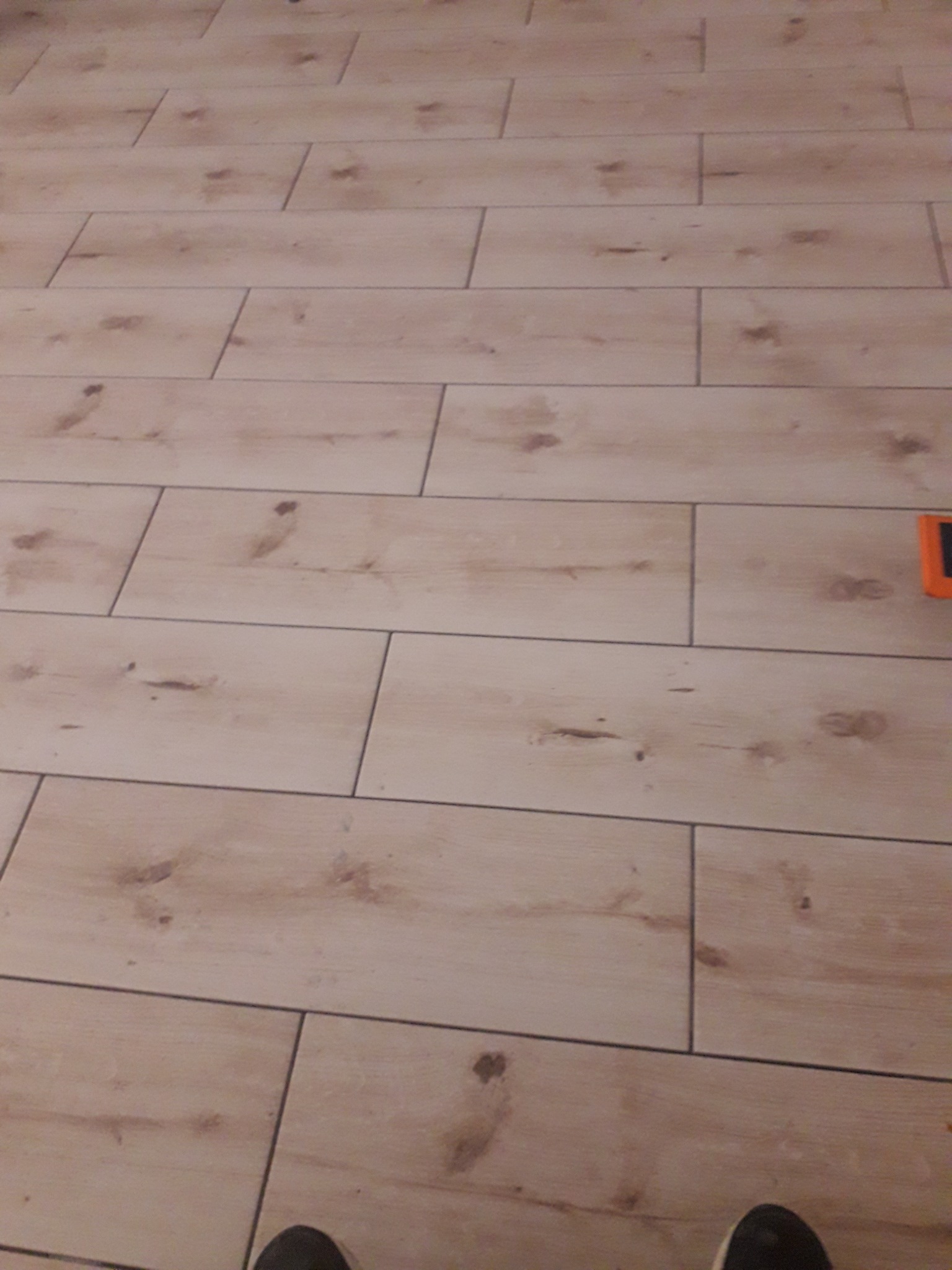 Jasne panele podłogowe imitujące drewno, ułożone w regularny wzór, z widocznym fragmentem buta w dolnej części kadru i pomarańczowym narzędziem w prawym górnym rogu.