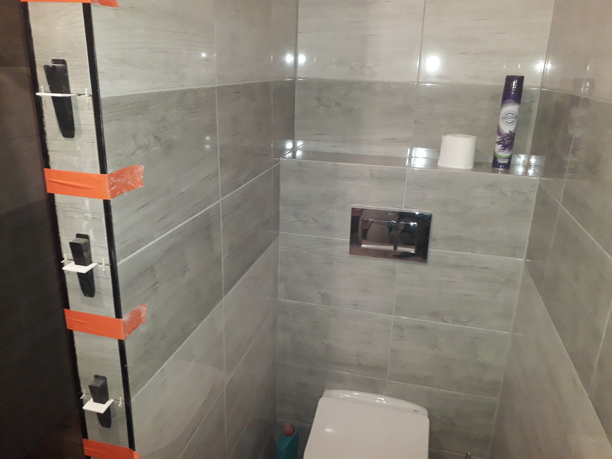 Montaż szarych płytek imitujących drewno w łazience, widoczne klipsy poziomujące i taśma malarska, częściowo zamontowana toaleta, dozownik mydła i odświeżacz powietrza.