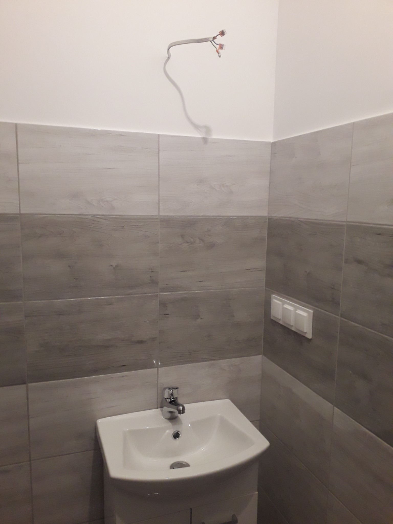 Umywalka narożna z szafką w łazience wyłożonej szaro-białymi płytkami imitującymi drewno, widoczne wystające przewody elektryczne nad umywalką i potrójny włącznik światła na ścianie.