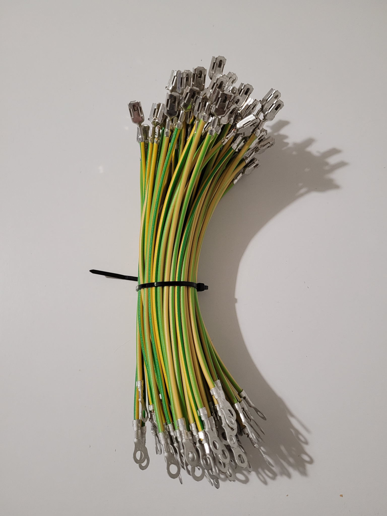 Wiązka przewodów elektrycznych w izolacji żółto-zielonej, z metalowymi końcówkami oczkowymi, spięta czarnym zaciskiem kablowym na białym tle.