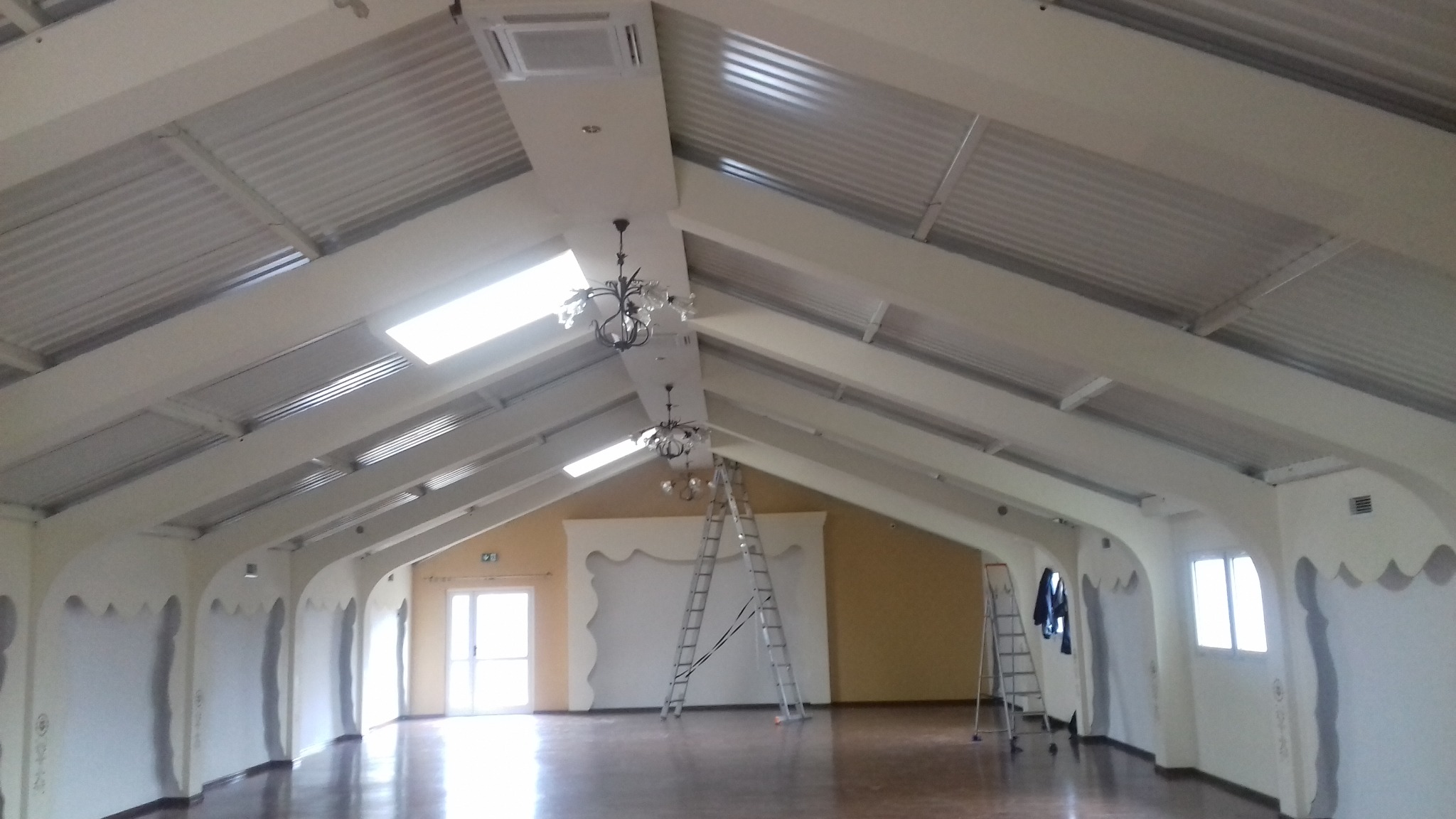 Przestronne wnętrze sali z wysokim, skośnym sufitem z widocznymi belkami, oświetlone przez okna dachowe i wiszące żyrandole. Ściany w trakcie szpachlowania, z widocznymi śladami po pracach. Dwie...