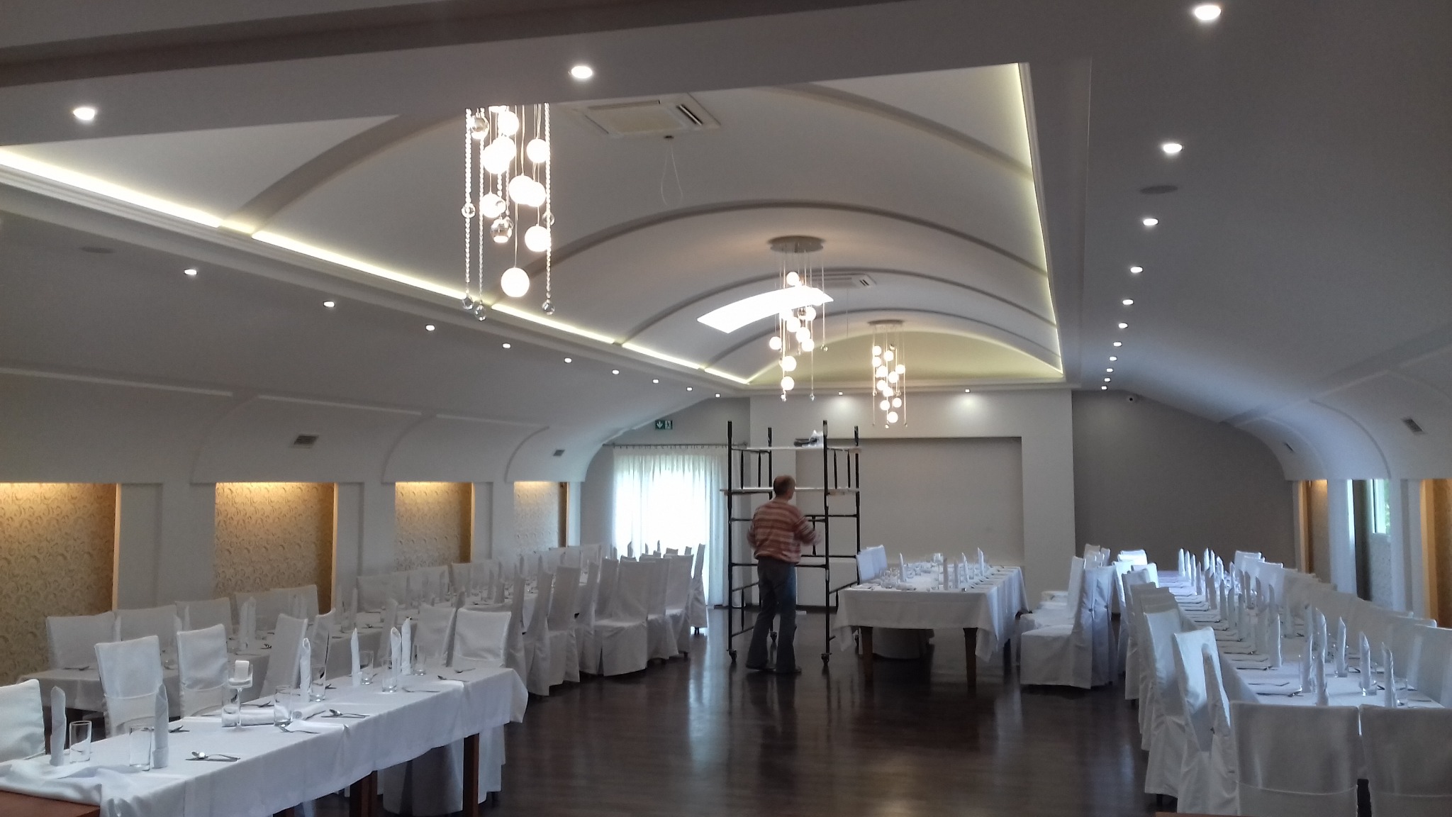 Sala bankietowa z dekoracyjnymi sufitami podwieszanymi i oświetleniem LED, przygotowana na przyjęcie, pracownik ustawia dekoracje.