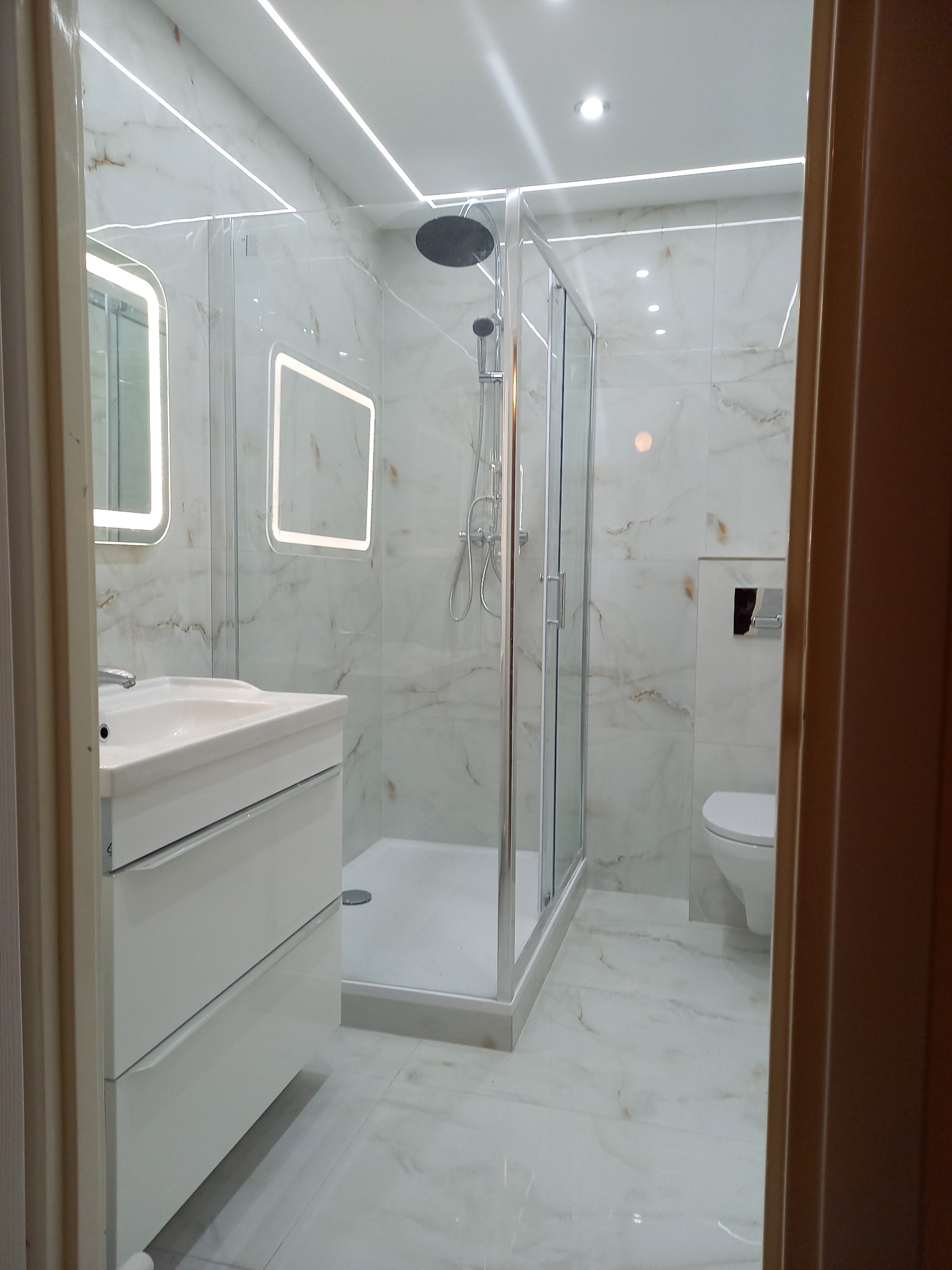 Wykończona łazienka z marmurowymi płytkami, kabiną prysznicową typu walk-in z deszczownicą, podświetlanymi lustrami i wiszącą toaletą.