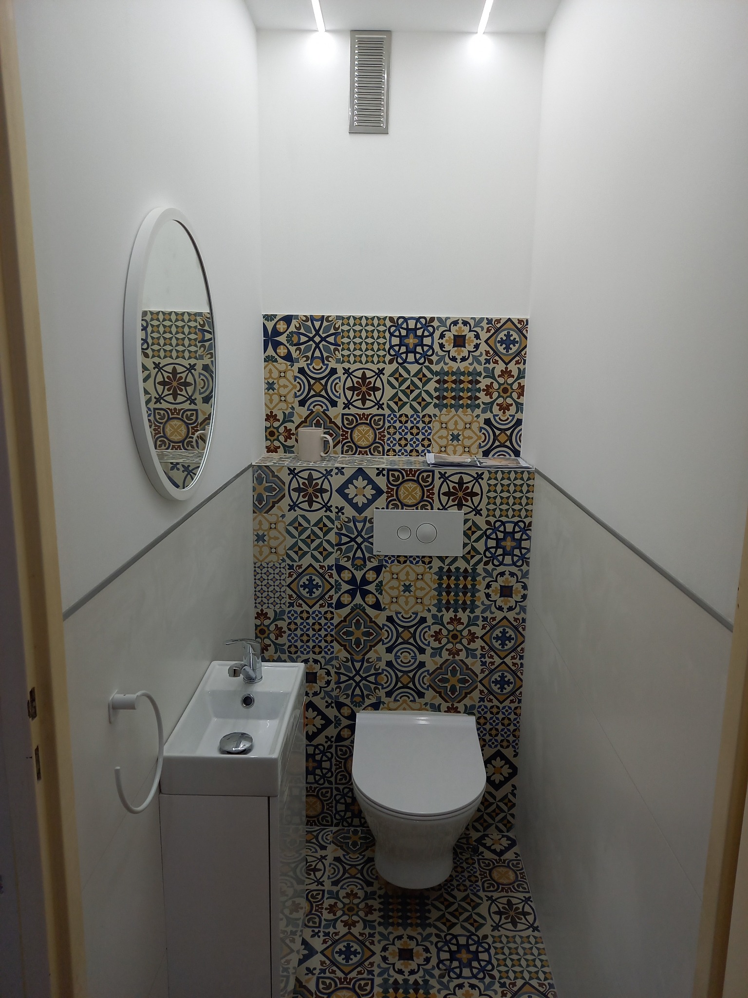 Wąska toaleta z wzorzystymi płytkami na ścianie i podłodze, biała miska ustępowa podwieszana, mała umywalka z szafką, okrągłe lustro, oświetlenie LED w suficie.