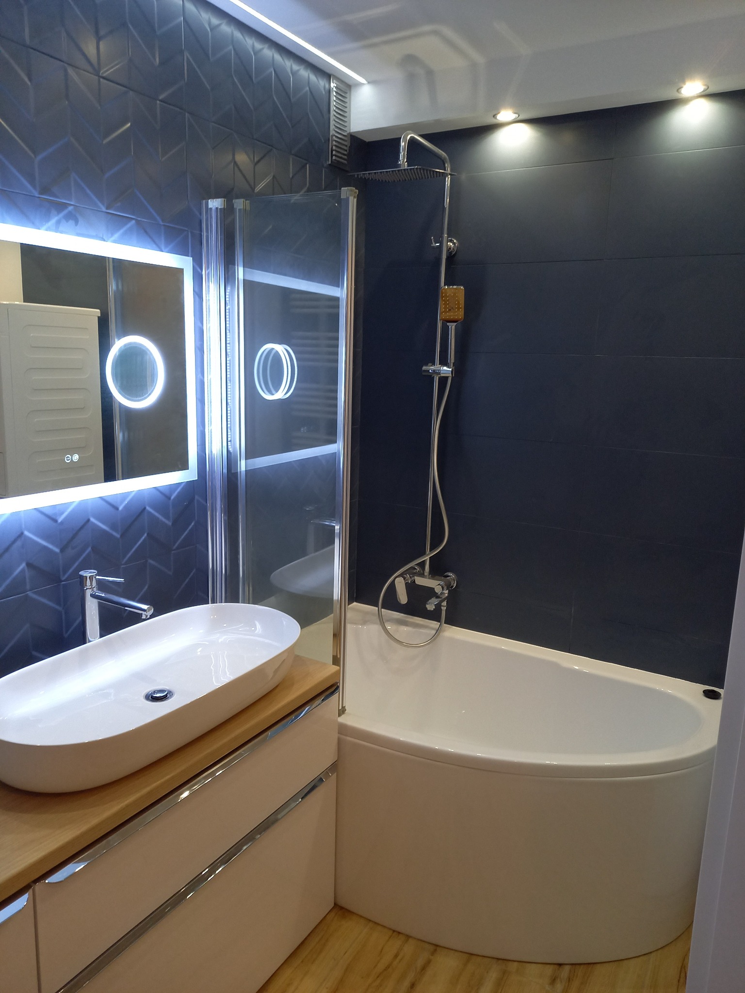 Nowoczesna łazienka z ciemnymi, wzorzystymi płytkami, owalną umywalką nablatową na jasnym blacie, podświetlanym lustrem i narożną wanną z parawanem.