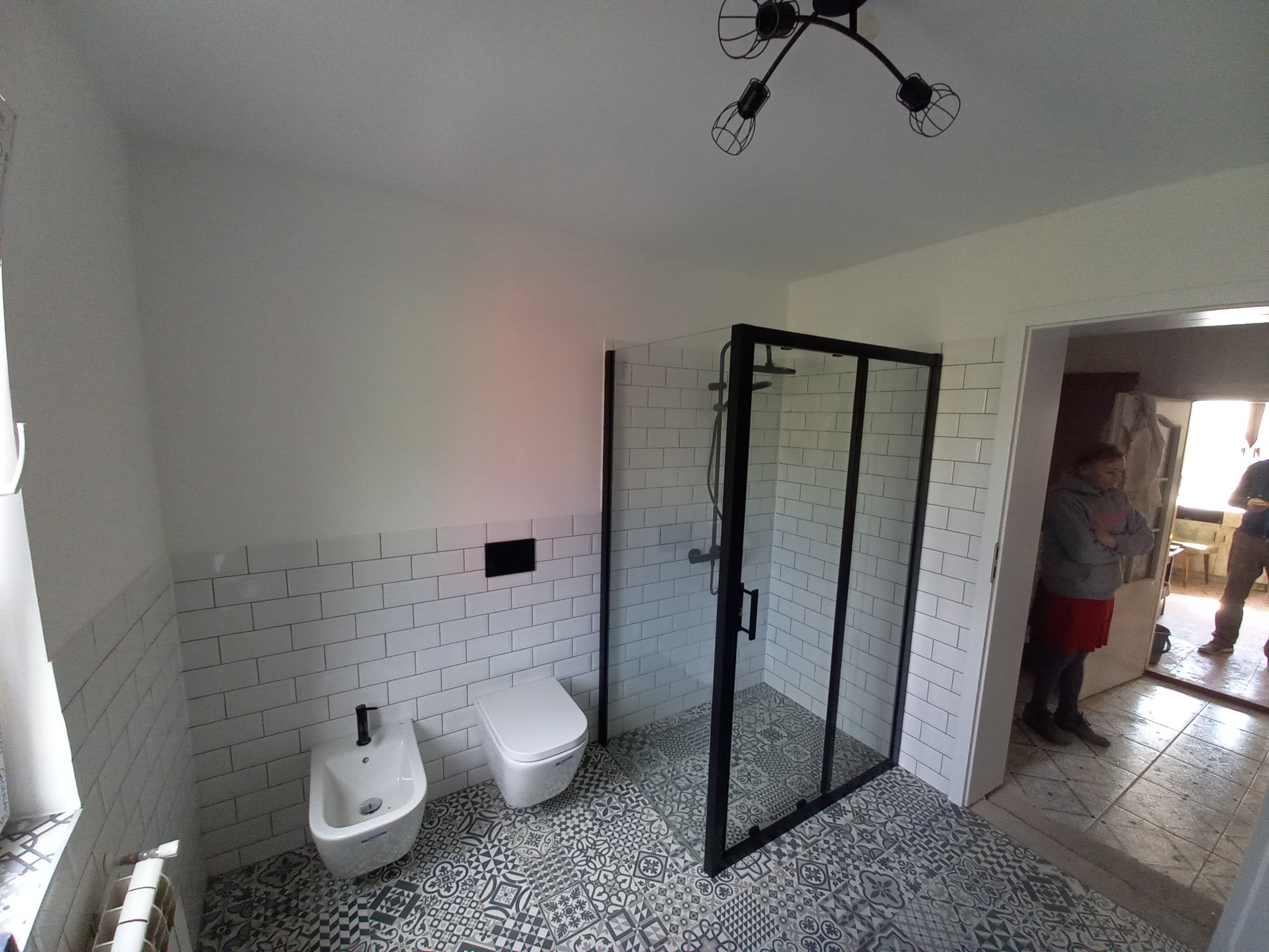 Nowoczesna łazienka po remoncie: kabina prysznicowa z czarną ramą, białe płytki na ścianach, wzorzysta podłoga, bidet i toaleta w minimalistycznym stylu, widoczna lampa sufitowa z żarówkami Edison.