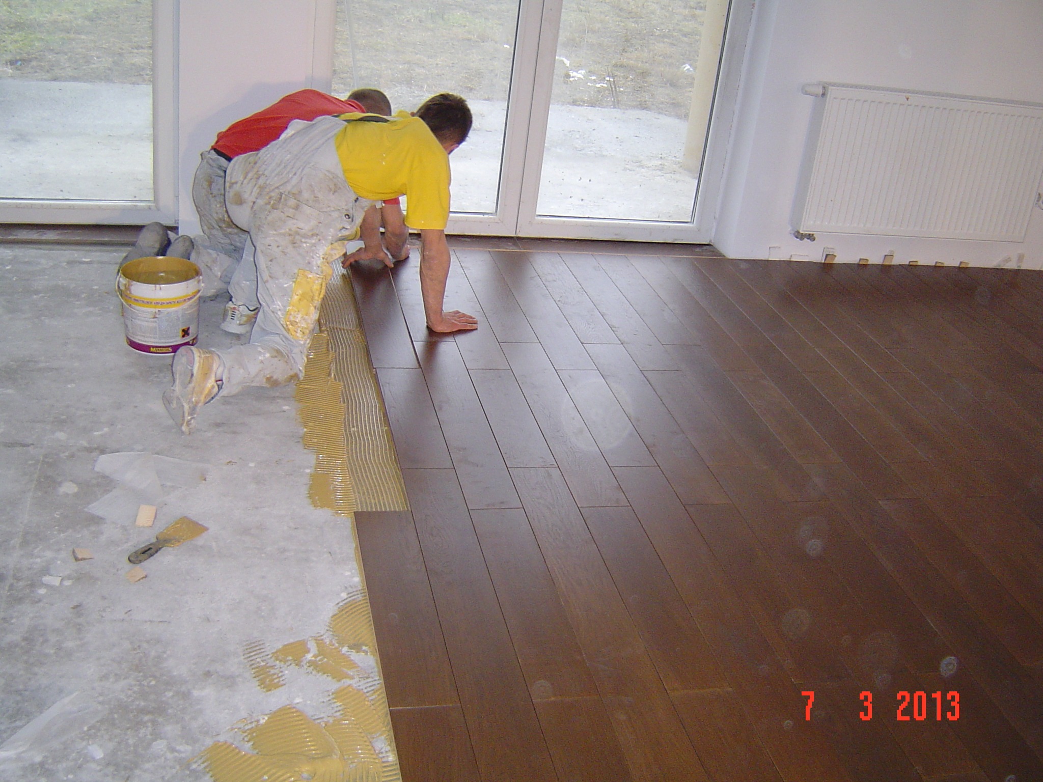 Dwóch mężczyzn w roboczych kombinezonach układa ciemne panele podłogowe na klej, widoczny fragment surowej podłogi i narzędzia, okno balkonowe w tle.