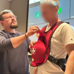 Instruktor demonstruje użycie kamizelki ratunkowej z symulatorem tonięcia podczas kursu pierwszej pomocy, w tle ekran z wideokonferencją.