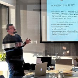 BHP - Szkolenia Nadzór Doradztwo - Prelegent gestykuluje podczas prezentacji o warunkach bezpieczeństwa i higieny pracy w pomieszczeniach, wyświetlanej na projektorze. Na pierwszym planie widoczne laptopy, szklanki z wodą i kawa...