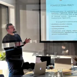 Prelegent gestykuluje podczas prezentacji o warunkach bezpieczeństwa i higieny pracy w pomieszczeniach, wyświetlanej na projektorze. Na pierwszym planie widoczne laptopy, szklanki z wodą i kawa...