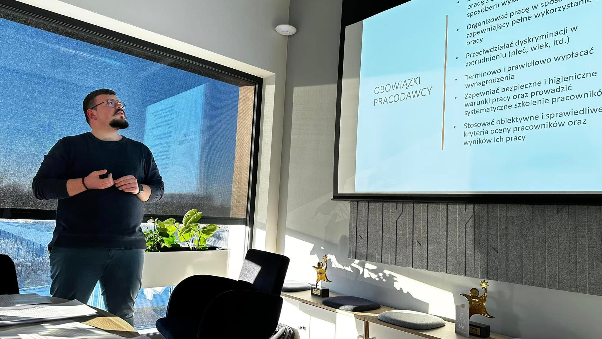 Prelegent w okularach prezentuje obowiązki pracodawcy na ekranie projektora, w tle okno z widokiem na budynek, na pierwszym planie stoliki i nagrody.