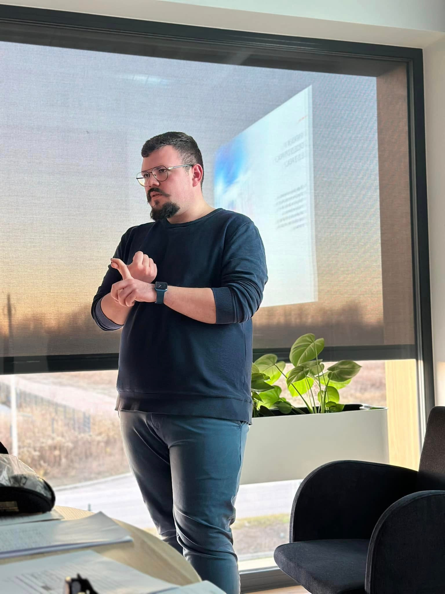 Mężczyzna w okularach i z brodą gestykuluje podczas prezentacji przed oknem z widokiem na krajobraz, na stole dokumenty i akcesoria biurowe.