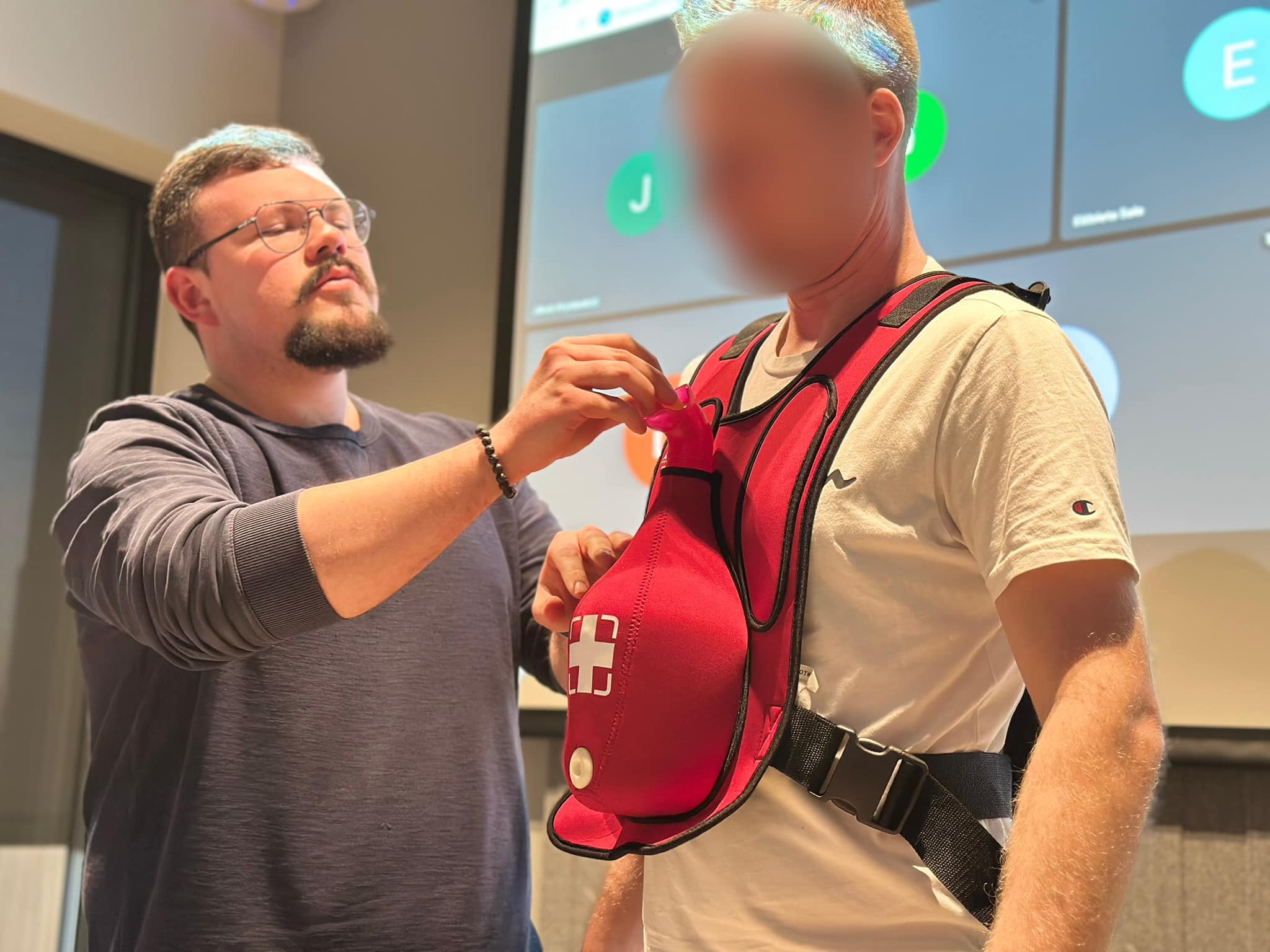 Instruktor demonstruje użycie kamizelki ratunkowej z symulatorem tonięcia podczas kursu pierwszej pomocy, w tle ekran z wideokonferencją.