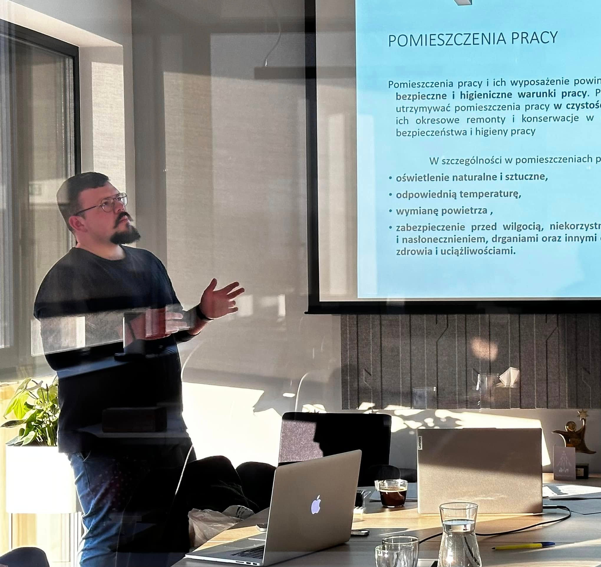 Prelegent gestykuluje podczas prezentacji o warunkach bezpieczeństwa i higieny pracy w pomieszczeniach, wyświetlanej na projektorze. Na pierwszym planie widoczne laptopy, szklanki z wodą i kawa...