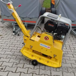 Żółta zagęszczarka gruntu marki Dro-Masz Professional Line na kostce brukowej w Krzyżanowie, z widocznym silnikiem spalinowym i przyczepami w tle.